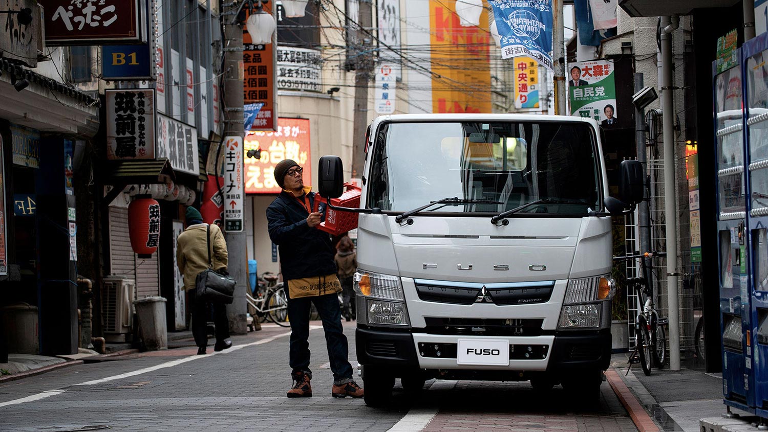 Camión Fuso 413 Blanco, mostrando la parte frontal del camión estacionado en un barrio chino mientras un operador carga bandejas de bebestibles