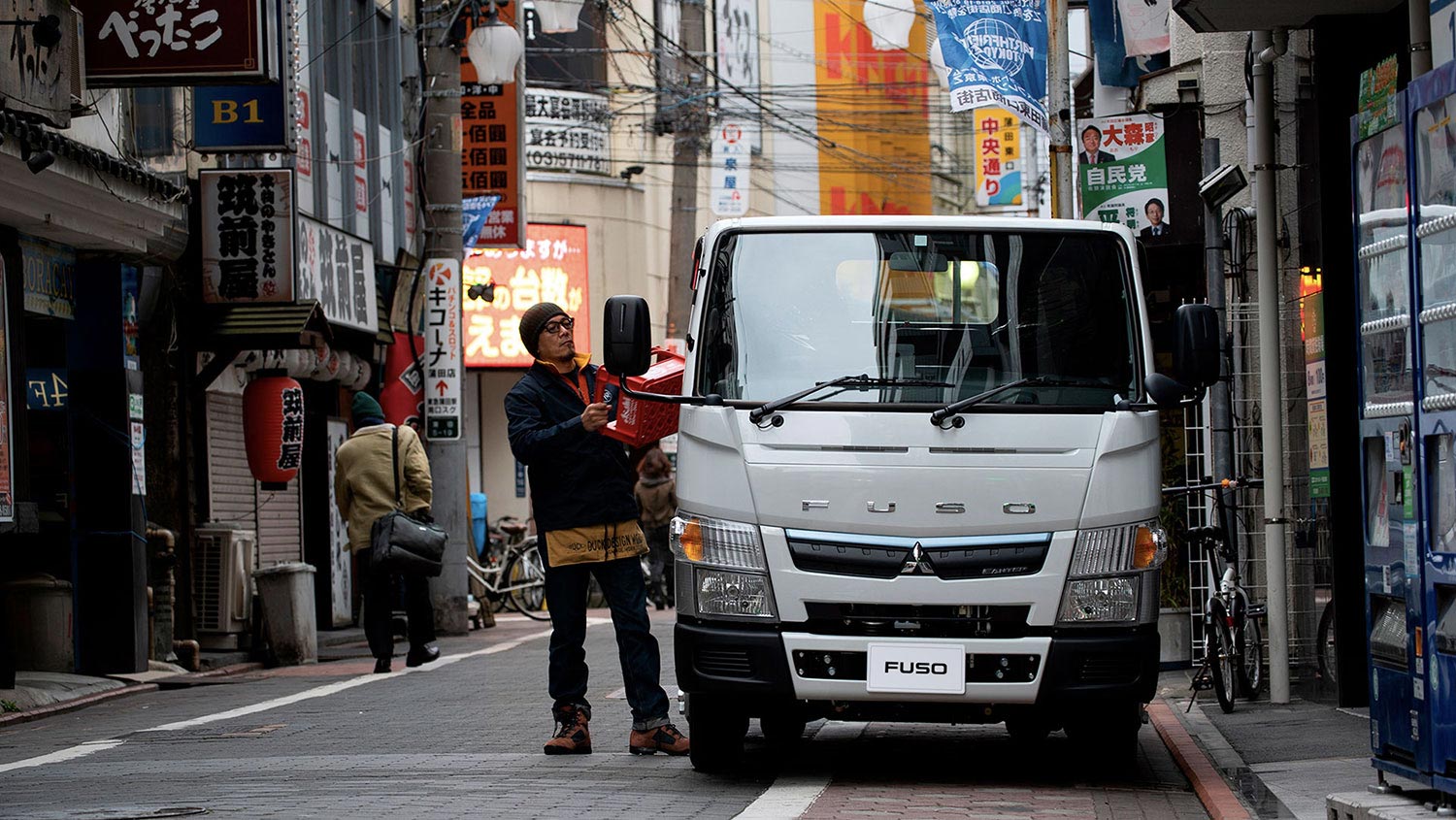 Camion Fuso 413 Blanco, mostrando la parte frontal del camion estacionado en un barrio chino mientras un operador carga bandejas de bebestibles