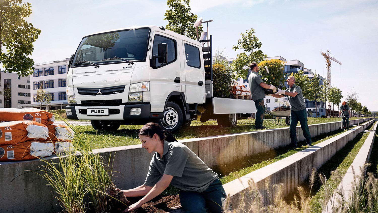 Camion Fuso 715 DC Blanco, estacionado alrededor de edificios mientras trabajadores colocan cargamento de plantas y sacos de tierra en el camion