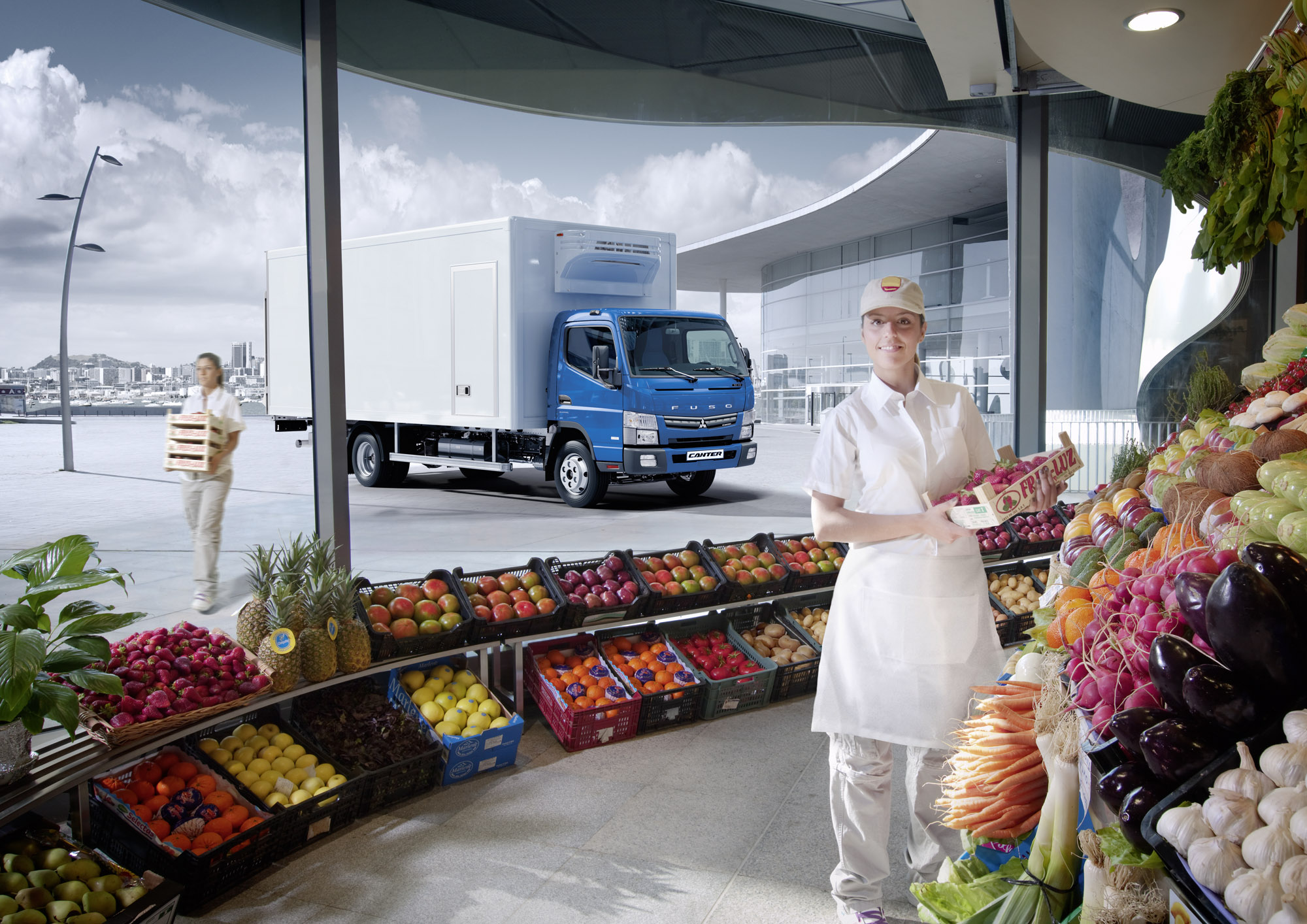 Trabajadora sostiene una caja de frutillas en una tienda de frutas y verduras, y por delante de la tienda hay un camion fuso celeste estacionado para que descarguen caja de frutas